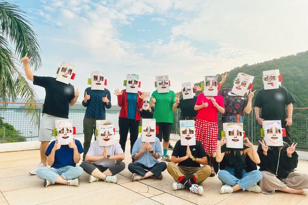 A group of workshop participants wearing decorated boxes on their heads, posing outdoors with a lake view