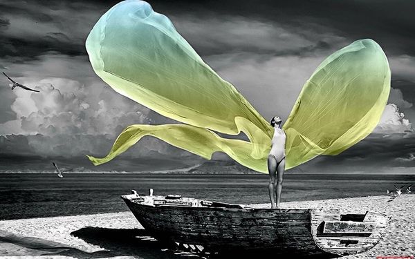 A woman standing on an old boat on a beach, with large yellow-green fabric wings spreading against a stormy sky